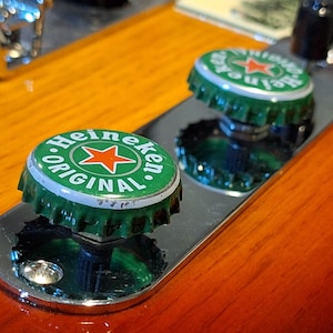 Op de afbeelding: Close-up van twee Heineken-flesdoppen met groene en witte branding, inclusief een rode ster. De doppen zijn bevestigd op een glanzend, zilveren metalen oppervlak, tegen een warme, houten achtergrond.