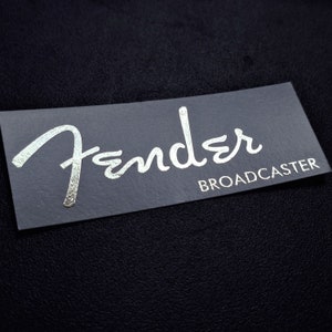 Peut inclure: Un logo Fender argenté sur un fond gris avec le mot "BROADCASTER" en noir en dessous.