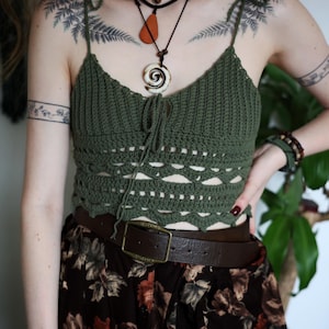 Könnte beinhalten: Ein grünes gehäkeltes Crop-Top mit Bindeträgern und einem dekorativen Lochmuster. Das Top wird mit einem Rock mit Blumenmuster und einem braunen Ledergürtel mit quadratischer Schnalle kombiniert. Eine mehrreihige Halskette und Armbänder vervollständigen den Look.