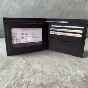 Könnte beinhalten: Geöffnete schwarze Ledergeldbörse mit mehreren Kartenfächern und einem transparenten Ausweisfach. Die Geldbörse enthält mehrere Karten und eine Visitenkarte mit Text.