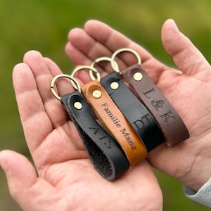 Op de afbeelding: Vier leren sleutelhangers in zwart, bruin en beige, elk met een goudkleurige ring. De sleutelhangers zijn gegraveerd met initialen en namen. De sleutelhangers worden in een open hand gehouden tegen een vage groene achtergrond.