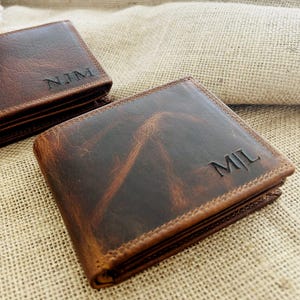 Könnte beinhalten: Zwei braune Ledergeldbörsen mit personalisierten Initialen. Die Geldbörsen sind rechteckig mit abgerundeten Ecken und einem genähten Rand. Das Leder hat ein abgenutztes Aussehen. Eine Geldbörse hat "NJM" und die andere "MIL" eingraviert.