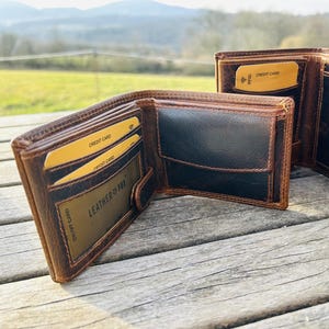 Puede incluir: Una cartera plegable de cuero marrón y negro con múltiples ranuras para tarjetas, un bolsillo para monedas con solapa y una ventana transparente. El interior incluye tarjetas amarillas y una tarjeta con el texto LEATHER FOX impreso.