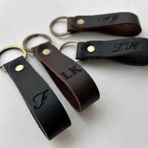 Puede incluir: Cuatro llaveros de cuero en colores marrón oscuro y negro, cada uno con un remache de metal dorado y una anilla circular. Las correas de cuero están grabadas con varias letras, incluyendo F, LK, ZH y AG en diferentes estilos de fuente.