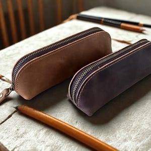 Pode incluir: Dois estojos de lápis de couro em tons de castanho e roxo, cada um com fecho de correr. Um lápis de madeira está em frente aos estojos. Os estojos são provavelmente para artigos de papelaria ou de arte.