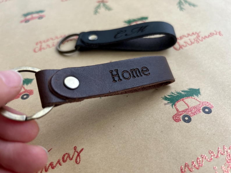 K&ouml;nnte beinhalten: Zwei Schl&uuml;sselanh&auml;nger aus Leder mit Metallringen und silberfarbenen Nieten. Ein Schl&uuml;sselanh&auml;nger ist dunkelbraun und mit dem Wort Home gepr&auml;gt, der andere ist schwarz und tr&auml;gt die eingepr&auml;gten Initialen C.M. auf dem Lederriemen.