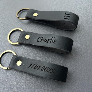 K&ouml;nnte beinhalten: Drei schwarze Schl&uuml;sselanh&auml;nger aus Leder mit messingfarbenen Metallringen und Nieten. Jeder Anh&auml;nger hat eine individuelle Gravur: oben die Buchstaben HF, in der Mitte der Name Charlie und unten das Datum 11.01.2022.