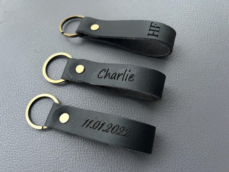 K&ouml;nnte beinhalten: Drei schwarze Schl&uuml;sselanh&auml;nger aus Leder mit messingfarbenen Metallringen und Nieten. Jeder Anh&auml;nger hat eine individuelle Gravur: oben die Buchstaben HF, in der Mitte der Name Charlie und unten das Datum 11.01.2022.