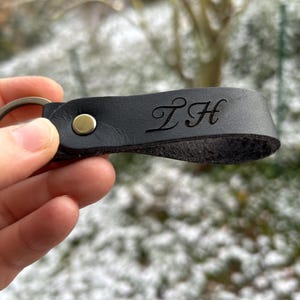 K&ouml;nnte beinhalten: Ein schwarzer Schl&uuml;sselanh&auml;nger aus Leder mit Schlaufendesign, das mit einer messingfarbenen Metallniete befestigt ist. Das Leder ist mit den stilisierten Gro&szlig;buchstaben ZH in Schreibschrift gepr&auml;gt. Ein runder Metallring ist am Ende befestigt.