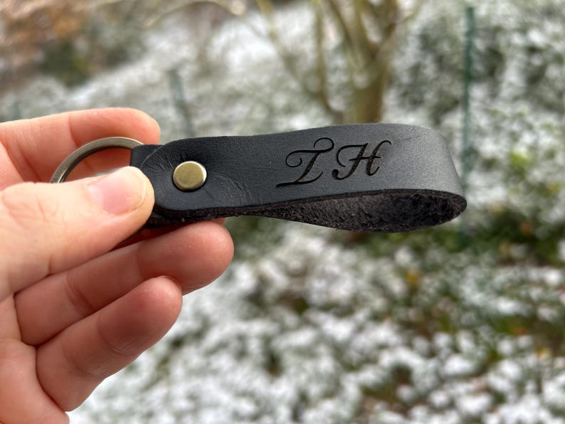 K&ouml;nnte beinhalten: Ein schwarzer Schl&uuml;sselanh&auml;nger aus Leder mit Schlaufendesign, das mit einer messingfarbenen Metallniete befestigt ist. Das Leder ist mit den stilisierten Gro&szlig;buchstaben ZH in Schreibschrift gepr&auml;gt. Ein runder Metallring ist am Ende befestigt.