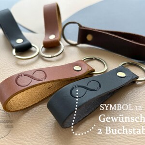 K&ouml;nnte beinhalten: F&uuml;nf Schl&uuml;sselanh&auml;nger aus Leder in Schwarz und Braun, jeweils mit Metallring und Niete. Zwei Anh&auml;nger sind mit einem Unendlichkeitssymbol gepr&auml;gt, das die Buchstaben ZE und ein kleines Herz enth&auml;lt. Text: SYMBOL 12 Gew&uuml;nschte 2 Buchstaben.