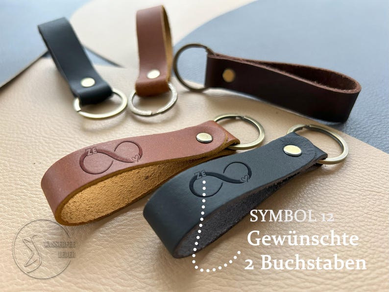 K&ouml;nnte beinhalten: F&uuml;nf Schl&uuml;sselanh&auml;nger aus Leder in Schwarz und Braun, jeweils mit Metallring und Niete. Zwei Anh&auml;nger sind mit einem Unendlichkeitssymbol gepr&auml;gt, das die Buchstaben ZE und ein kleines Herz enth&auml;lt. Text: SYMBOL 12 Gew&uuml;nschte 2 Buchstaben.