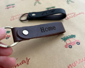 Könnte beinhalten: Zwei Schlüsselanhänger aus Leder mit Metallringen und silberfarbenen Nieten. Ein Schlüsselanhänger ist dunkelbraun und mit dem Wort Home geprägt, der andere ist schwarz und trägt die eingeprägten Initialen C.M. auf dem Lederriemen.