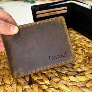 Könnte beinhalten: Ein braunes Lederportemonnaie mit dem Namen "Daniel" auf der Vorderseite. Das Portemonnaie ist geöffnet und zeigt Kreditkartenfächer und ein Geldfach. Das Leder hat einen Used-Look mit sichtbaren Nähten.