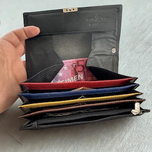 Könnte beinhalten: Schwarze Ledergeldbörse mit mehreren farbigen Fächern, darunter rot, blau und gelb. Die Geldbörse ist geöffnet und zeigt einen Fünf-Euro-Schein. Die Geldbörse hat einen Druckknopfverschluss und eine Reißverschlusstasche.