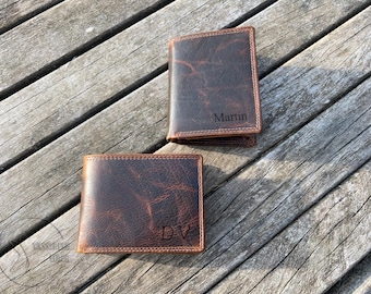 May include: Two dark brown leather wallets with a distressed texture and visible stitching along the edges. One wallet features the name Martin engraved on the front, while the other wallet displays the engraved initials DV on the bottom right corner.