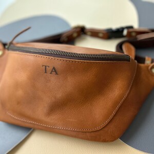 May include: A brown leather fanny pack with a zipper closure and a metal buckle. The initials "TA" are embossed on the front of the bag.