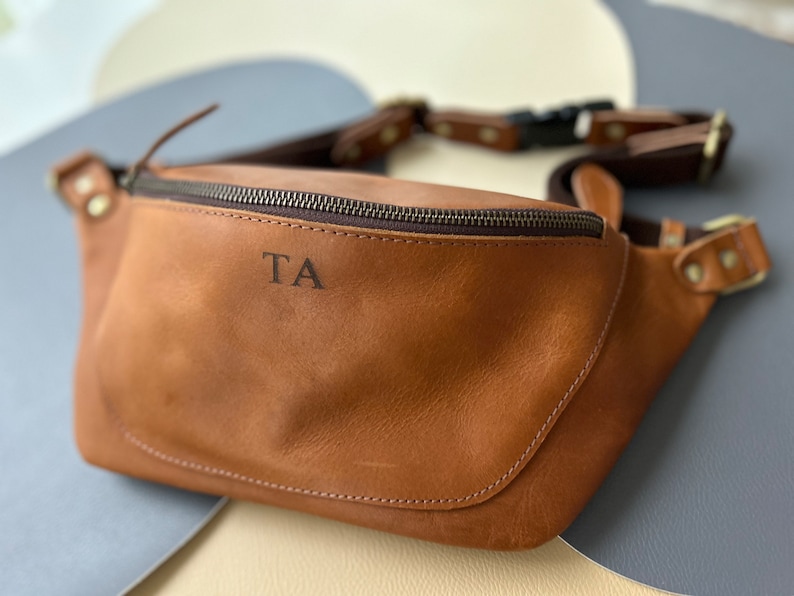 May include: A brown leather fanny pack with a zipper closure and a metal buckle. The initials "TA" are embossed on the front of the bag.