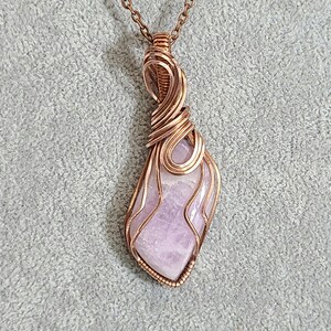 May include: A delicate copper wire wrapped pendant featuring a light purple gemstone. The pendant has a unique, asymmetrical design and is perfect for adding a touch of elegance to any outfit.