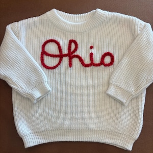 Puede incluir: Un suéter de punto color crema con la palabra "Ohio" bordada en rojo en el pecho. El suéter tiene mangas largas y un dobladillo y puños acanalados. La palabra "Ohio" está en cursiva.