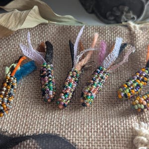 Könnte beinhalten: Fünf Perlenanhänger in Form von Maiskolben mit bunten Perlen und Schleifenakzenten liegen auf einer Jutefläche. Die Anhänger sind in verschiedenen Farben gehalten, darunter Blau, Orange und Grün. Ein schwarzes Band und ein dekorativer Kürbis befinden sich im Hintergrund.