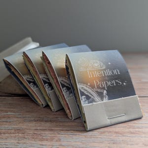 May include: A collection of Intention Papers with a celestial design, stacked on a wooden surface next to a white crystal. The papers have a silver and gold color scheme, with the words "Intention Papers" printed on the front.