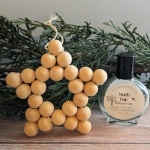 May include: A wooden star ornament made of small, round beads, with a string for hanging. Beside it is a clear glass bottle with a black cap, labeled "North Pole Diffuser Oil". The background features evergreen branches.