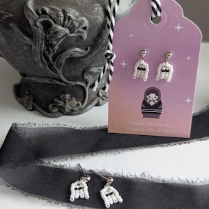 以下が含まれることがあります： 白いビーズと黒いアクセントで作られた手作りのゴーストイヤリング。ピンク色のタグに「Handmade jewelry, Magical Gifts & More!」の文字と白黒のコードが付いています。