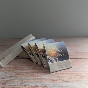 May include: A stack of Intention Papers with a decorative design and text on the cover, arranged on a wooden surface. A white, rectangular crystal is positioned to the left of the papers. The papers have a light blue and gold color scheme.