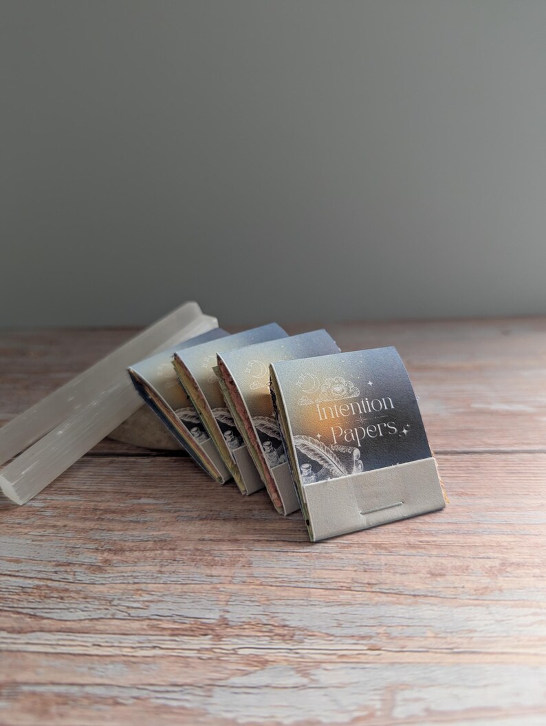 May include: A stack of Intention Papers with a decorative design and text on the cover, arranged on a wooden surface. A white, rectangular crystal is positioned to the left of the papers. The papers have a light blue and gold color scheme.