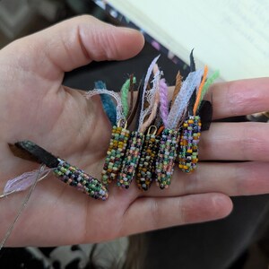 May include: A hand displays several beaded corn-shaped pendants. The pendants are crafted from small, colourful beads in shades of green, yellow, orange, and purple. Each pendant has a small loop at the top with colourful fabric and ribbon accents.