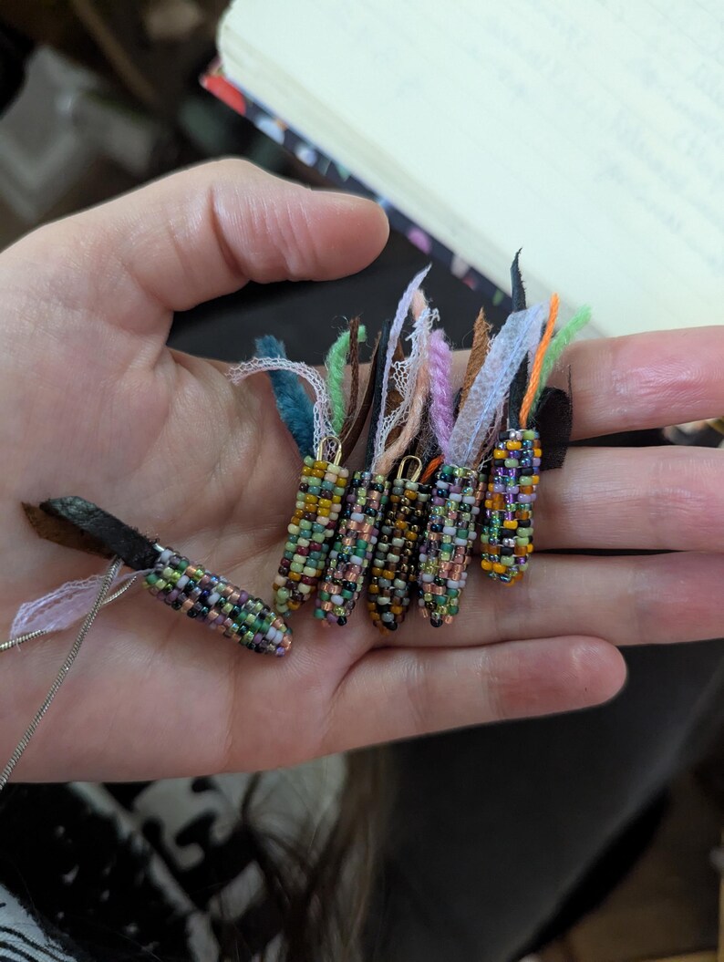 May include: A hand displays several beaded corn-shaped pendants. The pendants are crafted from small, colourful beads in shades of green, yellow, orange, and purple. Each pendant has a small loop at the top with colourful fabric and ribbon accents.