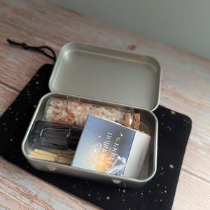 May include: An open silver metal tin filled with various items, including a glass bottle with pink crystals, a small glass vial, a black bottle, and a booklet labeled "Intention Papers." The tin sits on a black cloth with gold stars.