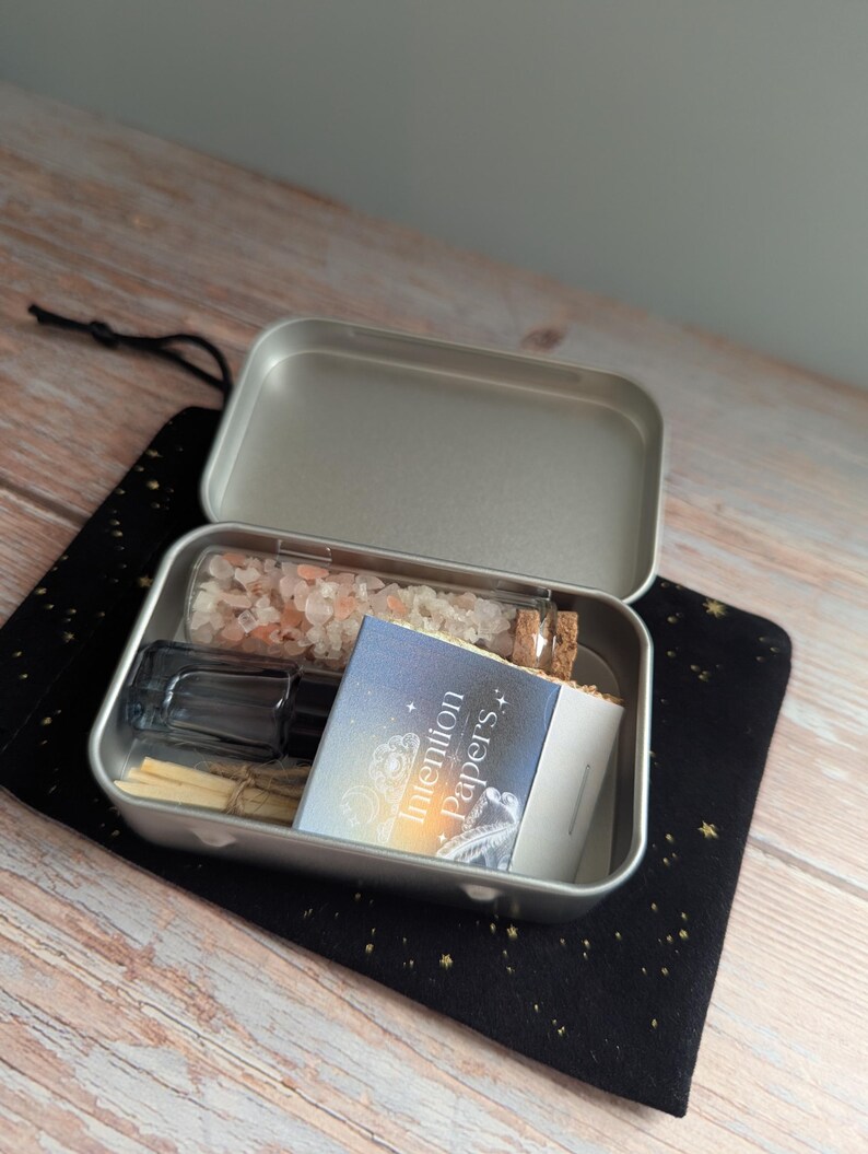 May include: An open silver metal tin filled with various items, including a glass bottle with pink crystals, a small glass vial, a black bottle, and a booklet labeled "Intention Papers." The tin sits on a black cloth with gold stars.