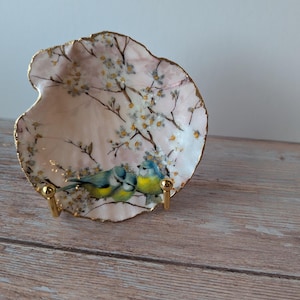 May include: A decorative, shell-shaped dish with a pink interior and gold trim. The dish features a painted design of blue and yellow birds perched on a flowering branch. The dish is supported by a gold-colored metal stand.
