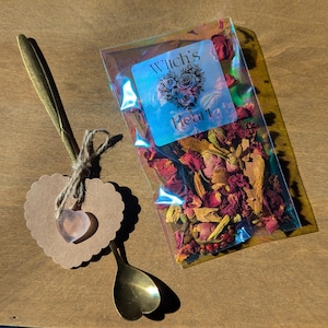 Könnte beinhalten: Ein kleiner transparenter Plastikbeutel gefüllt mit getrockneten Rosenblättern und anderen Kräutern. Der Beutel trägt ein Etikett mit der Aufschrift "Witch's Heart". Im Bild sind auch ein goldener Löffel und ein herzförmiges Etikett mit einem rosafarbenen herzförmigen Stein zu sehen.