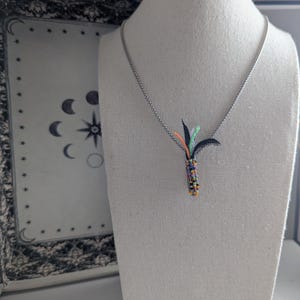 May include: A silver necklace with a pendant featuring a colourful beaded design and black, orange, and green accents. The pendant is shaped like a stylised plant. The necklace is displayed on a mannequin.