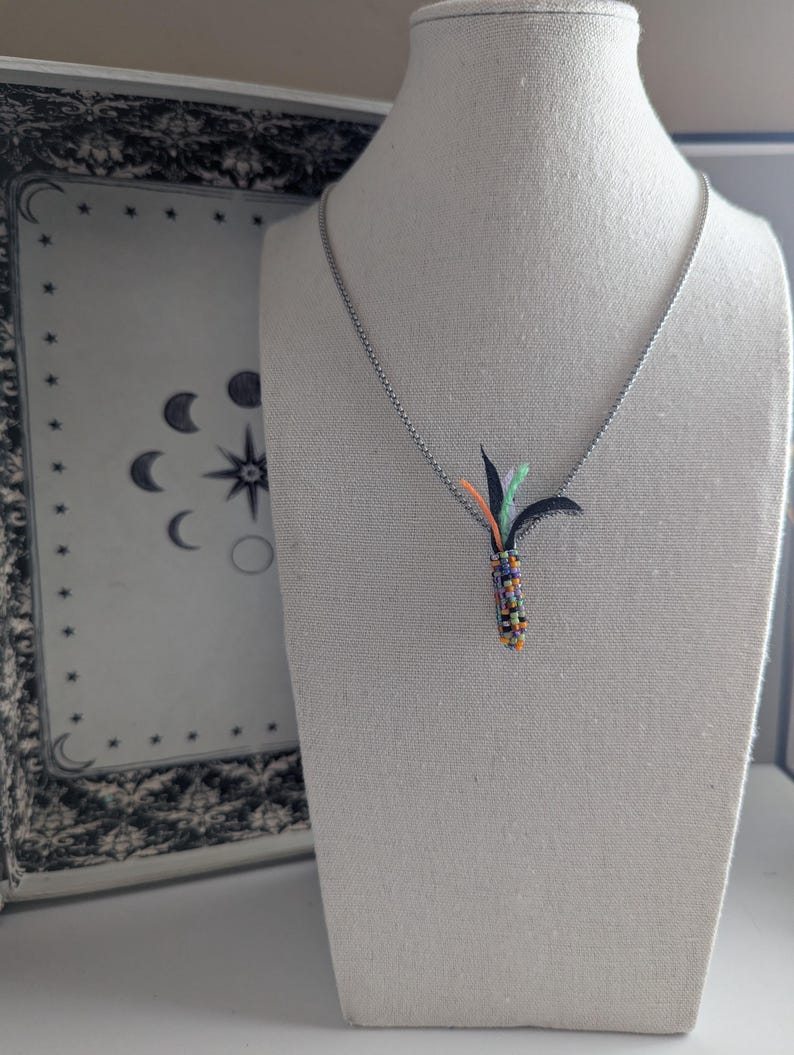 May include: A silver necklace with a pendant featuring a colourful beaded design and black, orange, and green accents. The pendant is shaped like a stylised plant. The necklace is displayed on a mannequin.