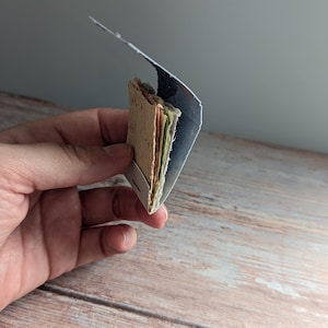 May include: A small, handmade paper booklet with colorful pages and a textured cover. The booklet is held in a hand, showcasing its compact size and unique, artisanal quality. The pages are visible, displaying a variety of colors.