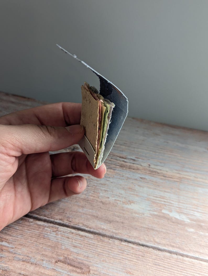 May include: A small, handmade paper booklet with colorful pages and a textured cover. The booklet is held in a hand, showcasing its compact size and unique, artisanal quality. The pages are visible, displaying a variety of colors.