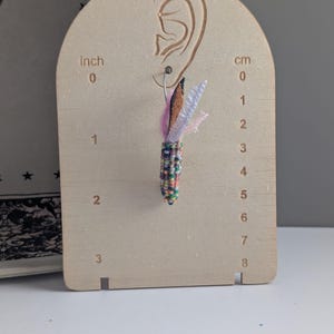 May include: A single beaded earring with a colourful design, featuring a brown, pink, and white feather-like accent. The earring is displayed on a wooden earring display board with cm measurements.