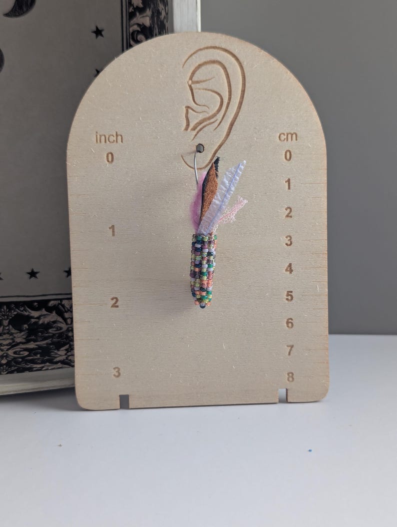 May include: A single beaded earring with a colourful design, featuring a brown, pink, and white feather-like accent. The earring is displayed on a wooden earring display board with cm measurements.