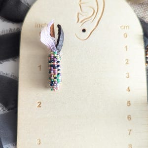 May include: A beaded earring with a colourful, cylindrical design, approximately 2.5 cm long, hangs from a black leather loop and white lace. The earring is displayed on a wooden earring display with an ear cutout and cm measurements.