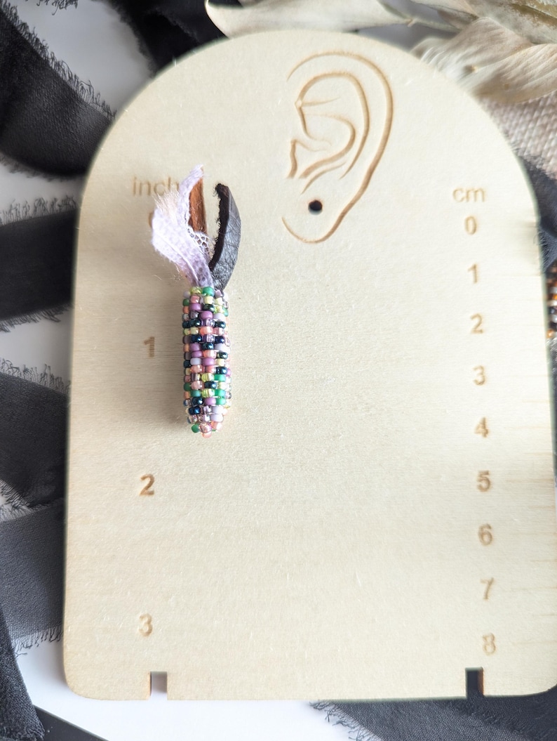 May include: A beaded earring with a colourful, cylindrical design, approximately 2.5 cm long, hangs from a black leather loop and white lace. The earring is displayed on a wooden earring display with an ear cutout and cm measurements.