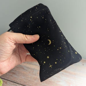 May include: Black drawstring bag with a celestial design. The bag features a pattern of gold stars and a crescent moon on a black background. The bag is held in a hand.