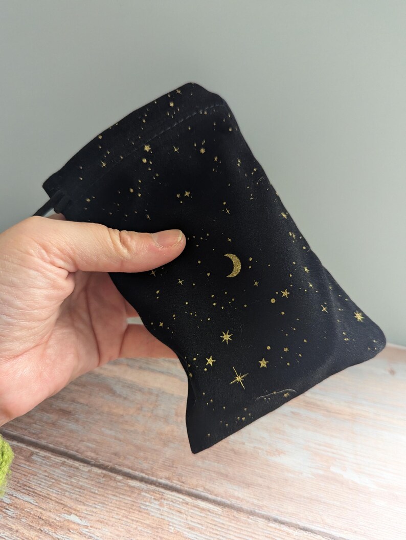 May include: Black drawstring bag with a celestial design. The bag features a pattern of gold stars and a crescent moon on a black background. The bag is held in a hand.