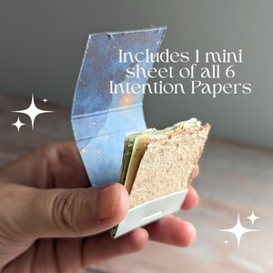 May include: A small, open container holding several sheets of paper. The container is white with a blue and gold patterned lid. The text "Includes 1 mini sheet of all 6 Intention Papers" is visible.