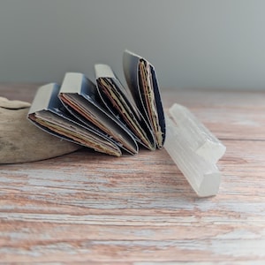 May include: A collection of handmade journals with blue covers and colorful pages, fanned out on a wooden surface. Two clear, crystalline selenite wands and a piece of driftwood are also present, creating a natural, artistic display.