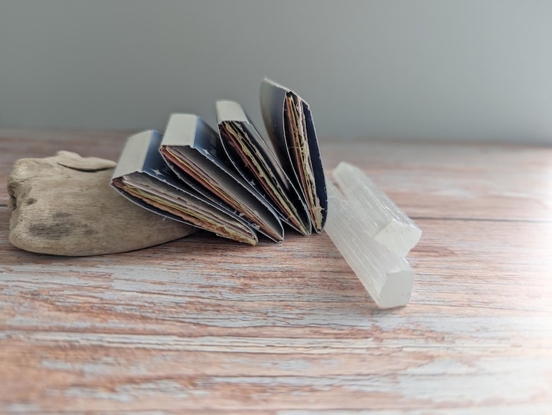 May include: A collection of handmade journals with blue covers and colorful pages, fanned out on a wooden surface. Two clear, crystalline selenite wands and a piece of driftwood are also present, creating a natural, artistic display.