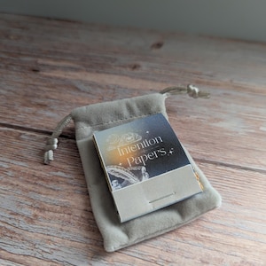 May include: A small, gray velvet pouch with a drawstring closure holds a booklet labeled "Intention Papers." The booklet has a starry night sky design and a feather illustration. The items rest on a wooden surface.